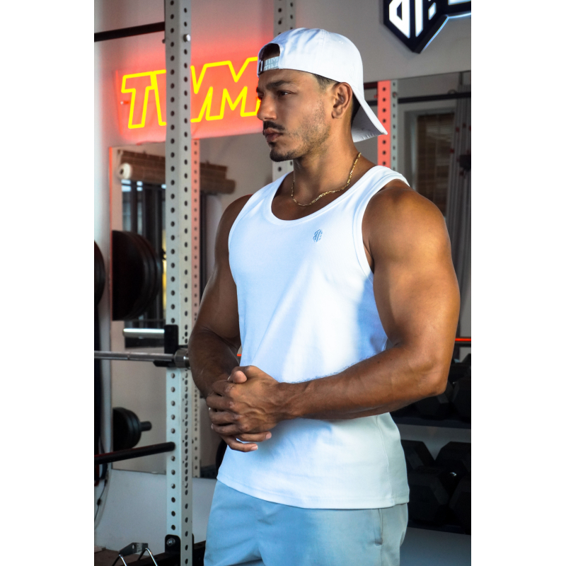 Homme musclé portant débardeur blanc ESSENTIAL, casquette arrière, chaîne dorée, en salle de sport avec poids et équipements au
