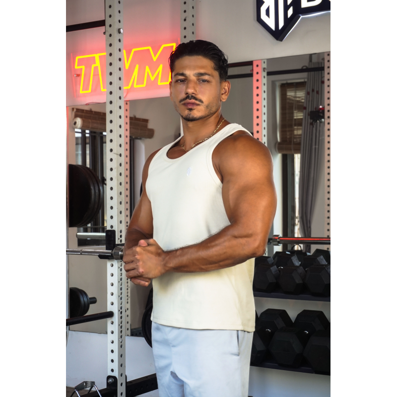 Homme musclé en débardeur clair dans une salle de sport, debout près d’un rack de poids et barres d’haltères.