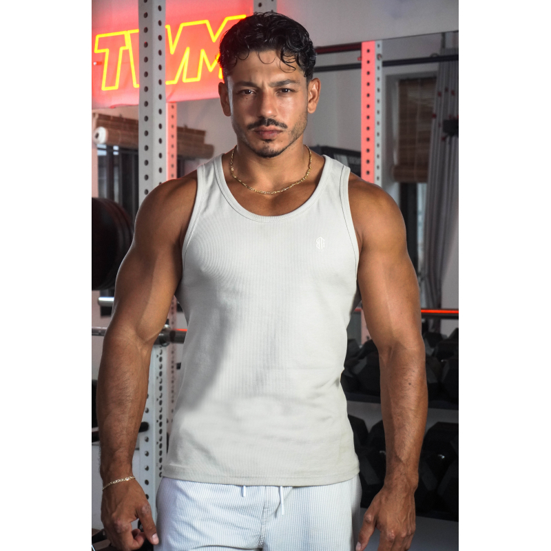 Homme musclé portant débardeur beige ESSENTIAL dans une salle de sport, avec collier doré et arrière gym.