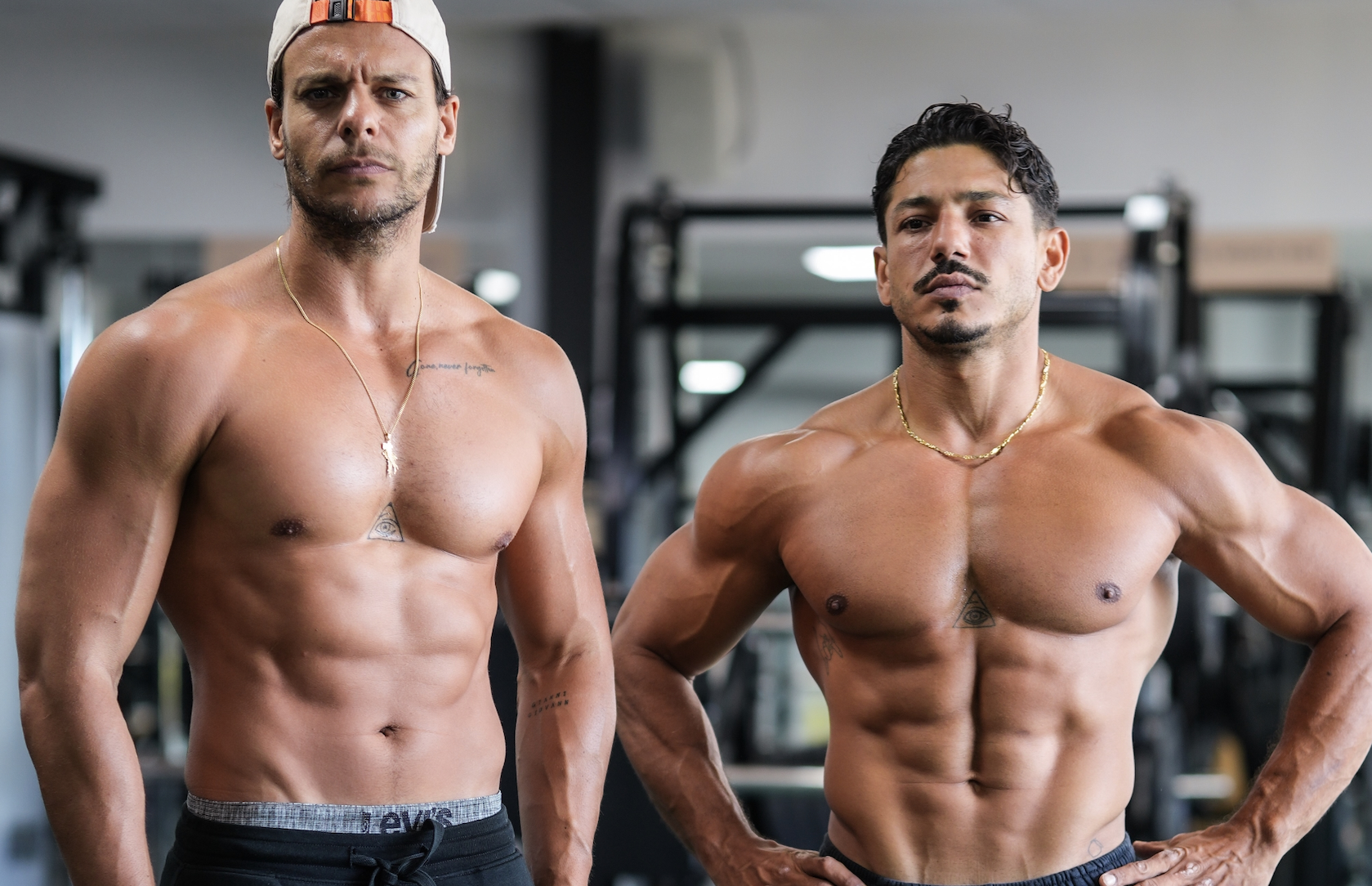 Deux coachs sportifs masculins, torse nu, posant dans une salle de gym avec machines en arrière-plan. L’un porte une casquette et un collier, l’autre chaîne dorée et tatouages visibles.