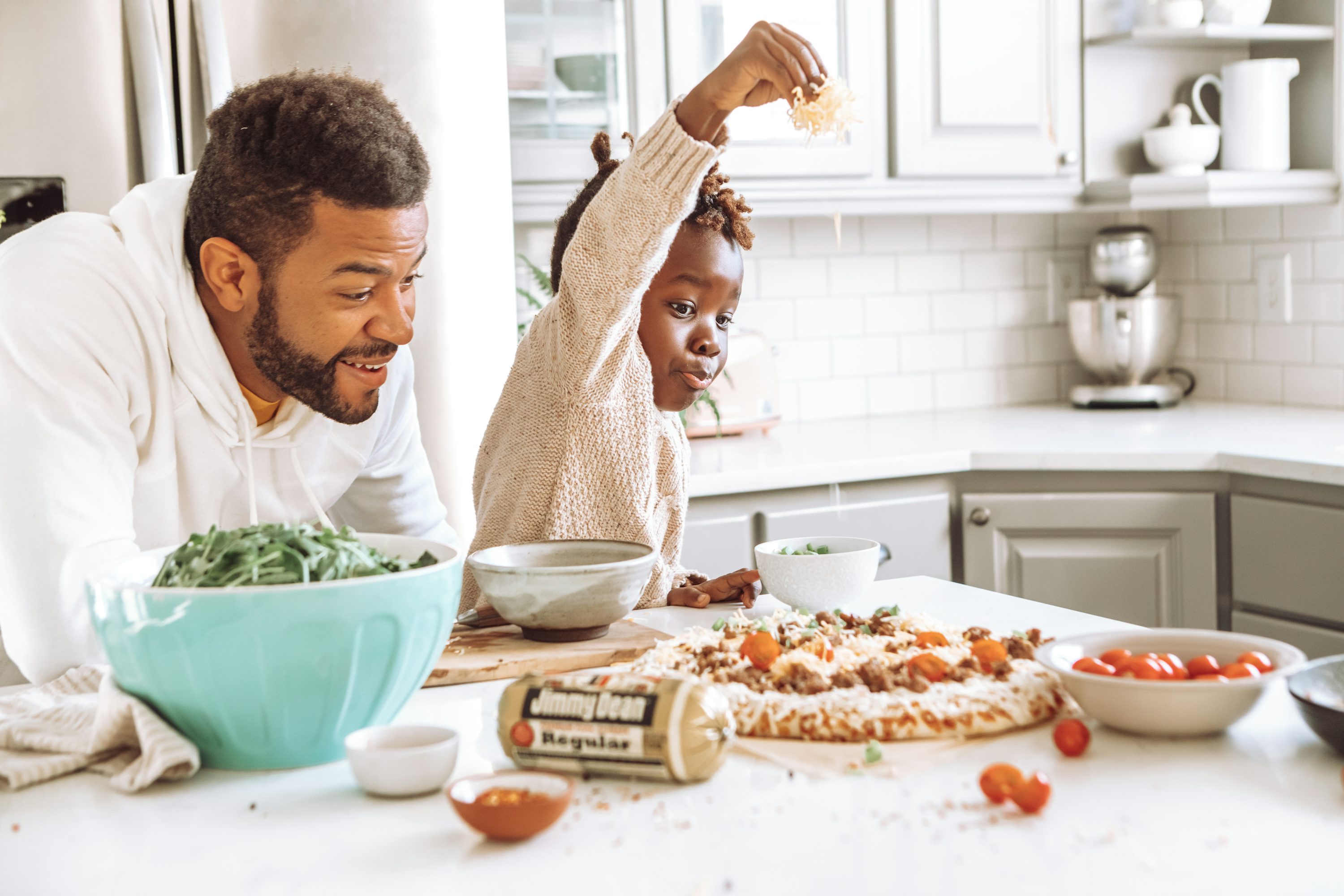 Un père et son enfant debout à une table de cuisine, entourés de nourriture: une pizza garnie, salade verte dans un grand bol bleu, tomates et assiettes, l’enfant lève les bras enjoué tandis que le père sourit.