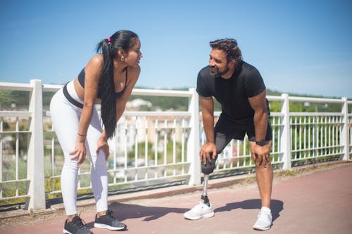 Deux coachs sportifs s’entraînent dehors sur une piste, échangent et rient, debout près d’une barrière blanche sous un ciel bleu.