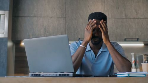 Homme devant un laptop, visiblement stressé, mains sur le front dans un bureau. Inclure le thème: comment gérer le stress au travail.