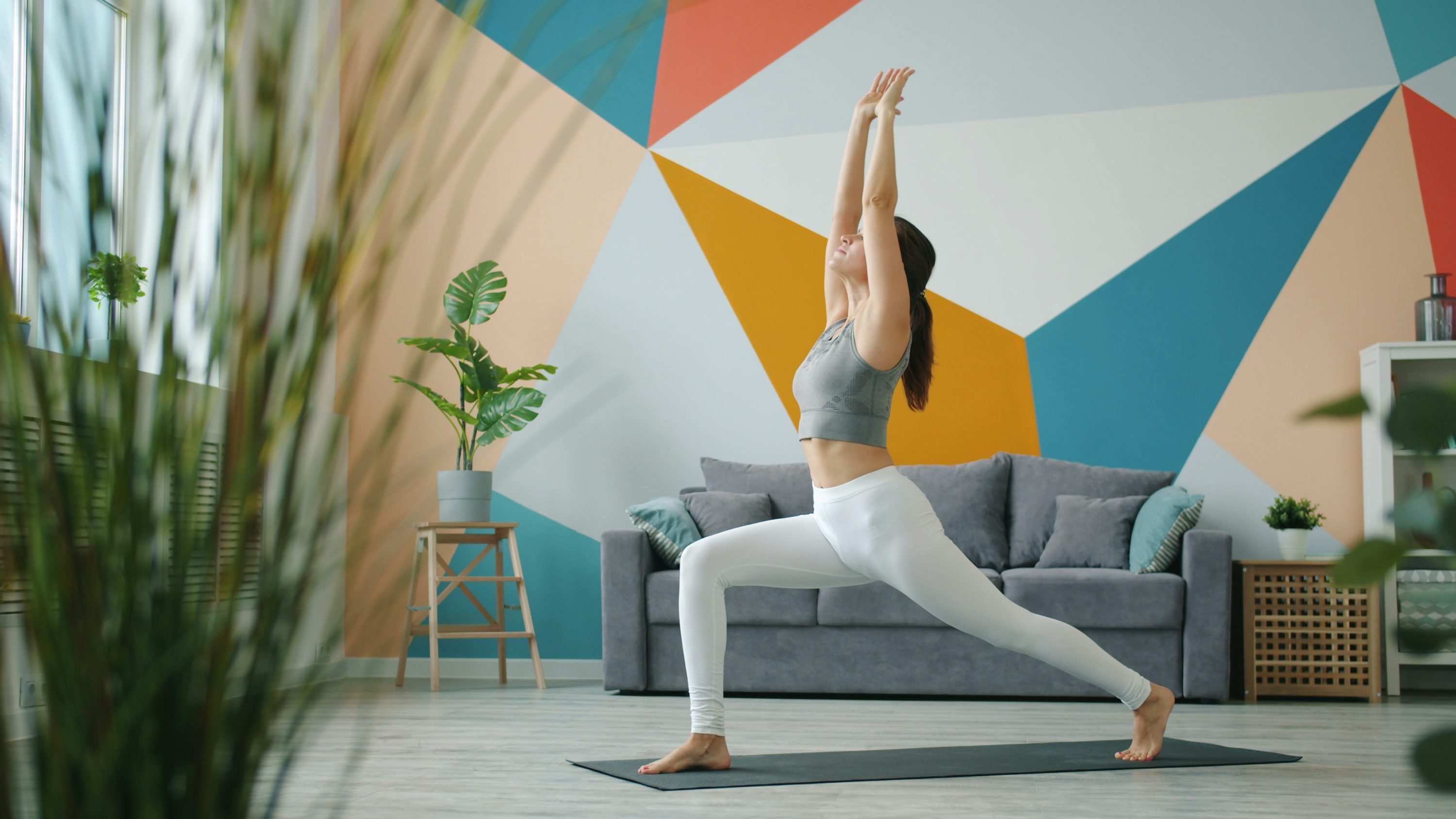 Jeune femme faisant une pose de yoga sur tapis, bras levés en l’air, dans un salon coloré et lumineux. intégré: comment gérer le stress au travail